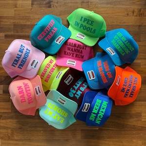 May include: A collection of twelve colorful foam trucker hats with mesh backs, each featuring unique text printed on the front. The hats come in various bright colors including pink, blue, green, orange, and yellow, with contrasting text colors.