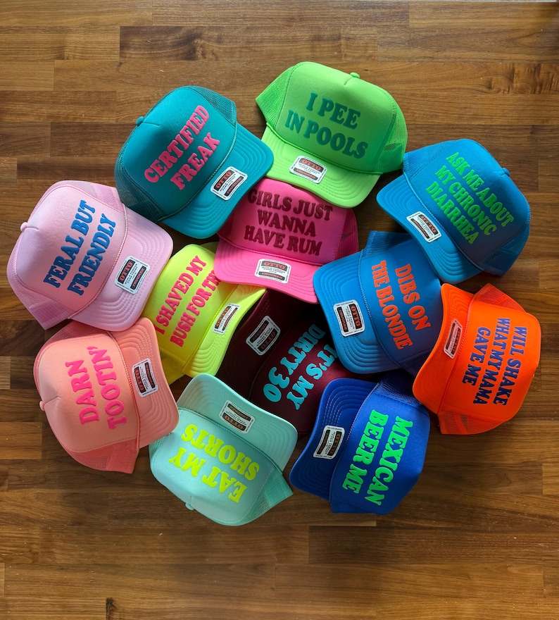 May include: A collection of twelve colorful foam trucker hats with mesh backs, each featuring unique text printed on the front. The hats come in various bright colors including pink, blue, green, orange, and yellow, with contrasting text colors.