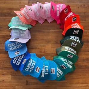 May include: A circular arrangement of multiple foam trucker hats in a spectrum of colors including pink, red, orange, green, and blue. Each hat features white block text with various phrases such as Tanned & Tipsy, Past My Bedtime, and Babe.