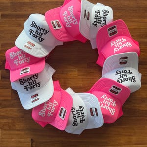 May include: A collection of trucker hats arranged in a circular pattern. The hats feature a white mesh back, a curved brim, and the text "Shorty hit Forty" in a bold, stylized font. The hats are primarily white and pink.