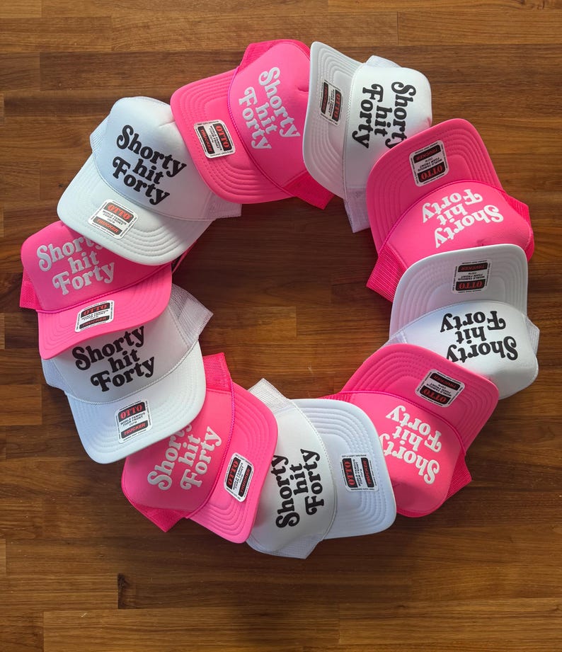 May include: A collection of trucker hats arranged in a circular pattern. The hats feature a white mesh back, a curved brim, and the text "Shorty hit Forty" in a bold, stylized font. The hats are primarily white and pink.