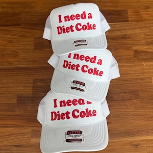 May include: Three white trucker hats stacked vertically, each featuring a foam front panel with the red text I need a Diet Coke printed in a bold font. Each hat includes a white mesh back, a curved brim, and an Otto brand sticker on the front.