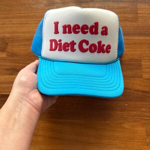 May include: A bright blue trucker hat featuring a white front panel with the red embroidered text I need a Diet Coke. The cap has a structured foam front, a curved brim, and a mesh back panel with a matching blue cord trim across the base.