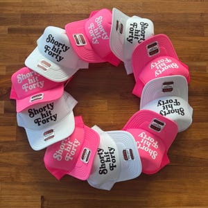 May include: A collection of trucker hats in a circular arrangement. The hats are in white and pink, with the text "Shorty Hit Forty" printed on the front. Each hat has a mesh back and a curved brim.