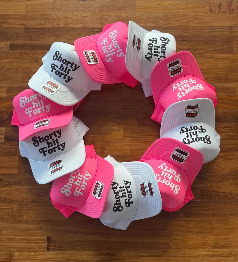 May include: A collection of trucker hats in a circular arrangement. The hats are in white and pink, with the text "Shorty Hit Forty" printed on the front. Each hat has a mesh back and a curved brim.