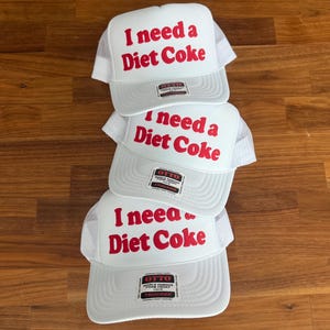 May include: Three white trucker hats stacked vertically, each featuring a foam front panel with the red text 'I need a Diet Coke' printed in a bold font. Each hat includes a white mesh back, a curved brim, and an Otto brand sticker on the front.
