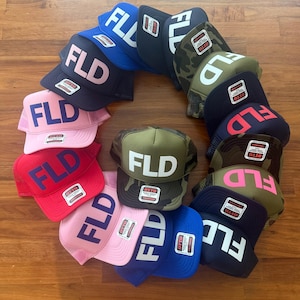 May include: A collection of twelve trucker hats arranged in a circle, each featuring the letters FLD on the front panel. The hats come in various colors including blue, black, pink, red, and camouflage patterns, all with Otto brand tags.