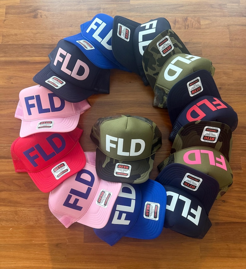 May include: A collection of twelve trucker hats arranged in a circle, each featuring the letters FLD on the front panel. The hats come in various colors including blue, black, pink, red, and camouflage patterns, all with Otto brand tags.