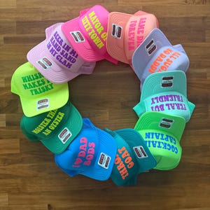 May include: A circular arrangement of eleven colorful trucker hats, each featuring a unique phrase in bold lettering. The hats come in various bright colors including neon green, pink, orange, blue, and teal, all displaying the Otto brand label.