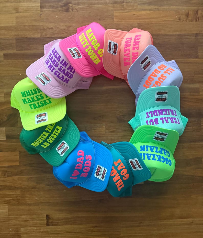 May include: A circular arrangement of eleven colorful trucker hats, each featuring a unique phrase in bold lettering. The hats come in various bright colors including neon green, pink, orange, blue, and teal, all displaying the Otto brand label.