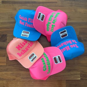 May include: Five foam front trucker hats in bright blue, neon pink, and peach colors. Each hat features a unique phrase in bold lettering: Catch Fly Not Feelin, Woke Up Sexy As Hell Again, Miss Behavin, Chaos Coordinator, and Too Hot To Behave.