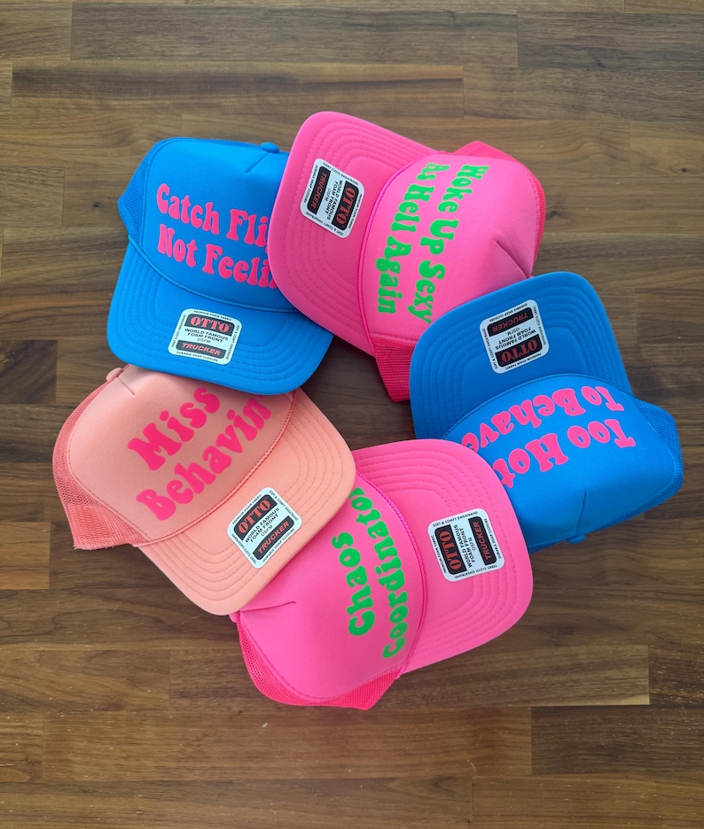 May include: Five foam front trucker hats in bright blue, neon pink, and peach colors. Each hat features a unique phrase in bold lettering: Catch Fly Not Feelin, Woke Up Sexy As Hell Again, Miss Behavin, Chaos Coordinator, and Too Hot To Behave.