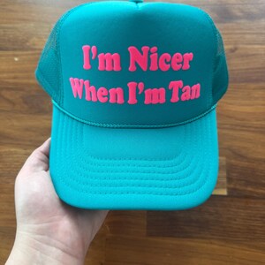 May include: A teal trucker hat featuring a mesh back and a solid front panel. The text "I'm Nicer When I'm Tan" is printed in bold, bright pink lettering across the center of the front panel. The hat includes a matching teal brim with stitching.