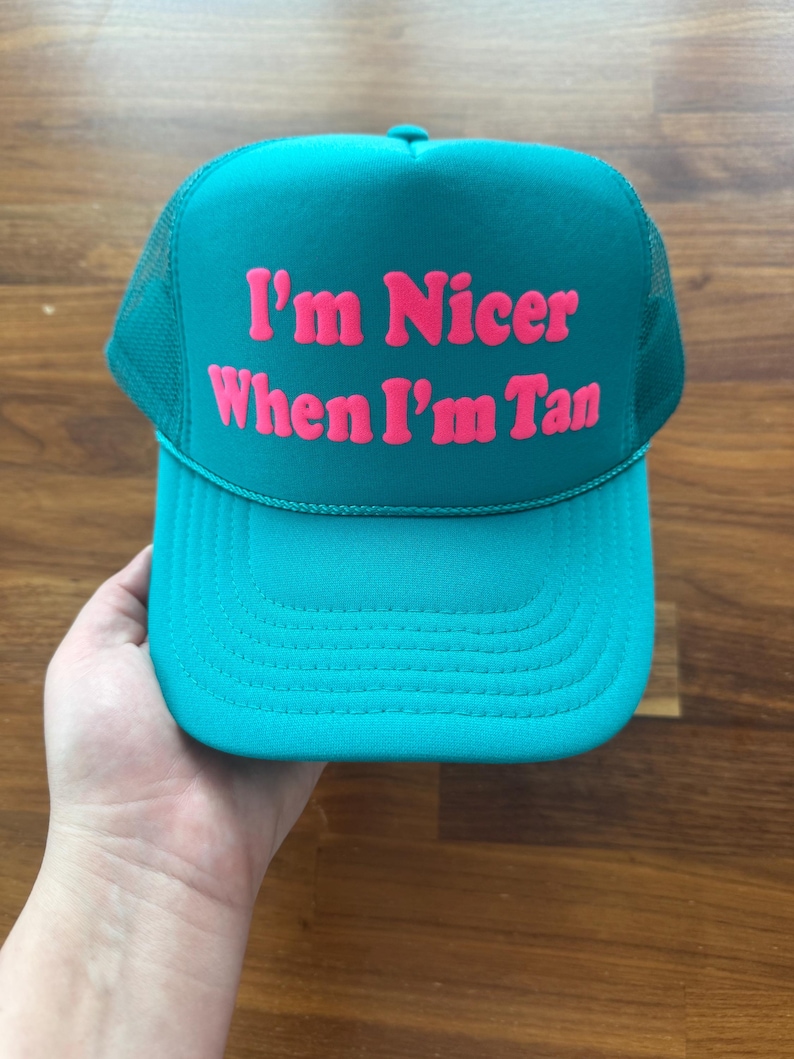 May include: A teal trucker hat featuring a mesh back and a solid front panel. The text "I'm Nicer When I'm Tan" is printed in bold, bright pink lettering across the center of the front panel. The hat includes a matching teal brim with stitching.