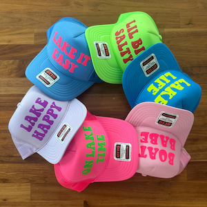May include: A collection of six foam front trucker hats in various bright colors including blue, lime green, pink, and lavender. Each hat features a unique phrase printed in a contrasting color, such as Lake It Easy, Lil Bi Salty, Lake Life, Lake Happy, On Lake Time, and Boat Babe.