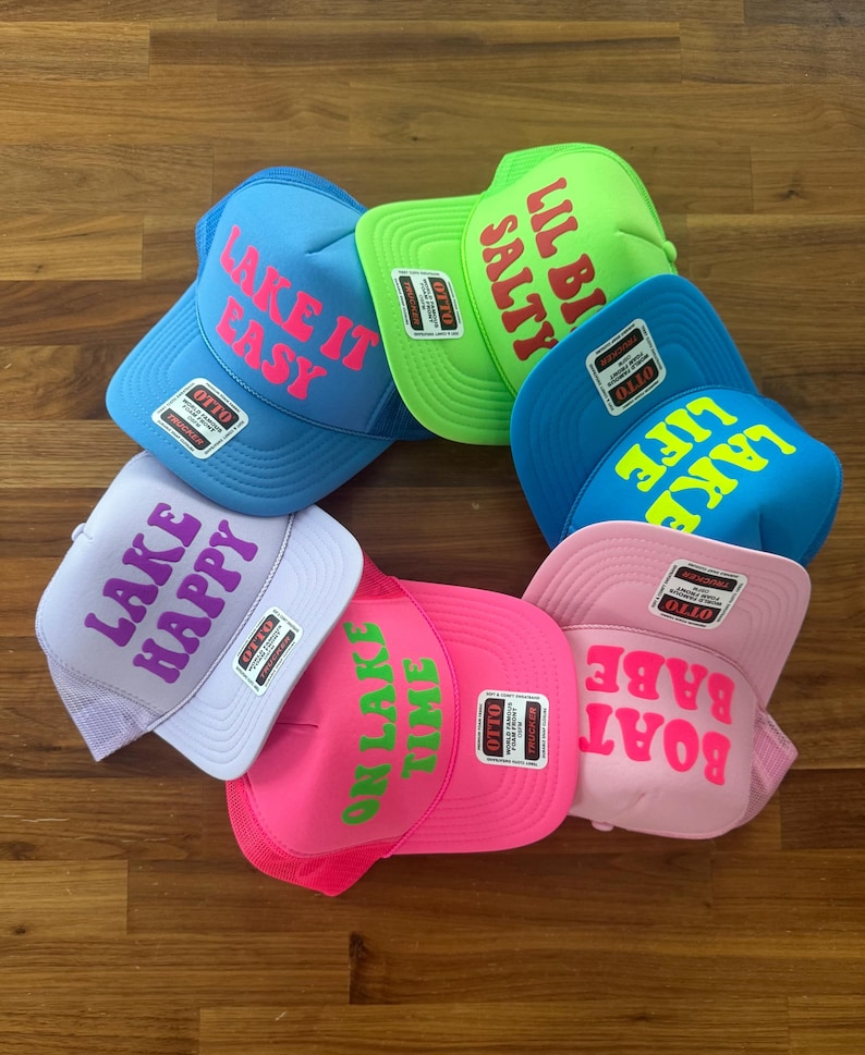 May include: A collection of six foam front trucker hats in various bright colors including blue, lime green, pink, and lavender. Each hat features a unique phrase printed in a contrasting color, such as Lake It Easy, Lil Bi Salty, Lake Life, Lake Happy, On Lake Time, and Boat Babe.