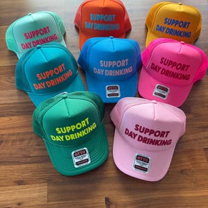 May include: Seven trucker hats in different colors with the text "Support Day Drinking" in yellow and red lettering. The hats are all made of mesh and have a velcro closure.