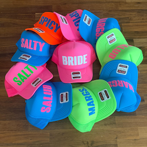 May include: A collection of neon-colored trucker hats featuring bold text including BRIDE, SALTY, SPICY, MARGS, and SALUD. Each hat has a structured foam front, a mesh back, and an Otto brand label on the brim. The hats are arranged in a cluster.