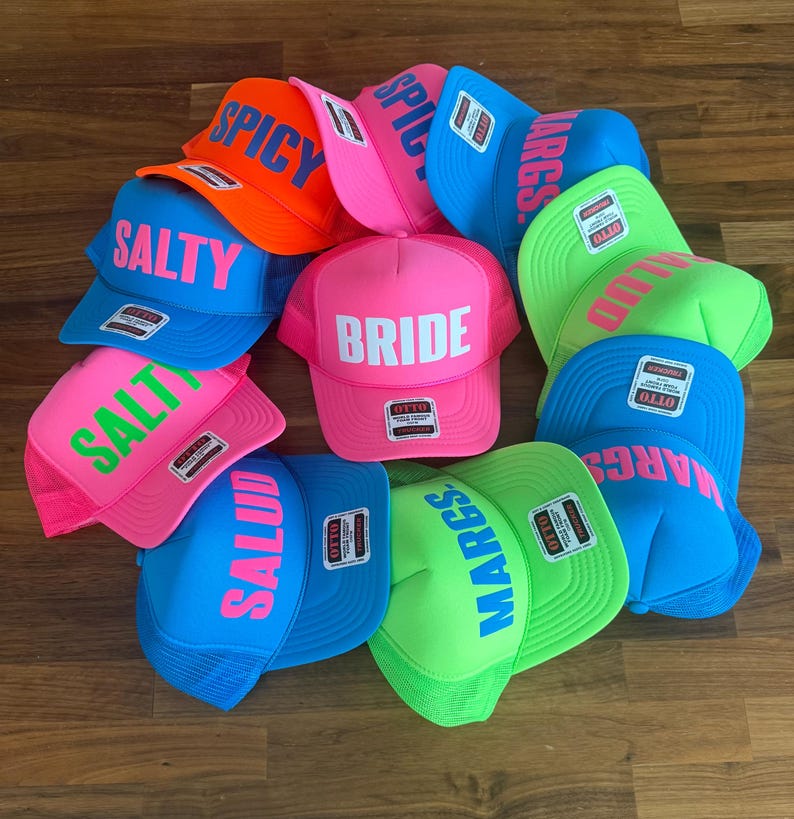 May include: A collection of neon-colored trucker hats featuring bold text including BRIDE, SALTY, SPICY, MARGS, and SALUD. Each hat has a structured foam front, a mesh back, and an Otto brand label on the brim. The hats are arranged in a cluster.