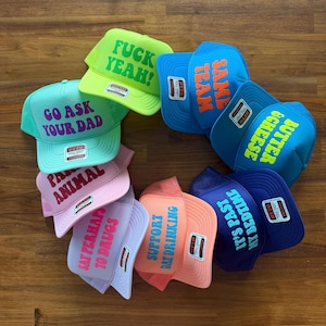 May include: A collection of eight colorful trucker hats arranged in a circle. Each hat features a unique phrase printed on the front, including FUCK YEAH, SAME TEAM, BUTTER & CHEESE, IT'S PAST MY BEDTIME, SUPPORT DAY DRINKING, SAY PERHAPS TO DRUGS, PARTY ANIMAL, and GO ASK YOUR DAD.