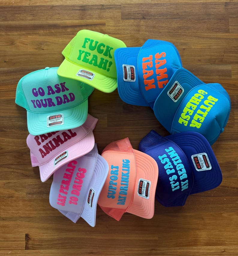 May include: A collection of eight colorful trucker hats arranged in a circle. Each hat features a unique phrase printed on the front, including FUCK YEAH, SAME TEAM, BUTTER & CHEESE, IT'S PAST MY BEDTIME, SUPPORT DAY DRINKING, SAY PERHAPS TO DRUGS, PARTY ANIMAL, and GO ASK YOUR DAD.