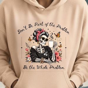 Könnte beinhalten: Ein beigefarbener Kapuzenpullover mit einem Grafikdruck eines Skeletts, das ein rotes Stirnband und eine Sonnenbrille trägt und eine Kaffeetasse hält. Der Text lautet, Don't Be Part of the Problem, Be the Whole Problem, umgeben von Schmetterlingen und Blumen.