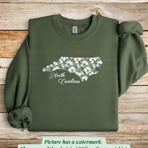 May include: An olive green sweatshirt featuring a floral North Carolina state outline. The state shape is filled with white flowers on a light blue background, with "North Carolina" in script below.