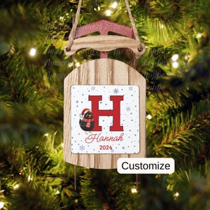 May include: A wooden Christmas ornament shaped like a sled with a red and white design. The ornament features a black and red bird with a Santa hat, a red letter H, and the name "Hannah" with the year "2024" below. The ornament is decorated with white snowflakes on a blue background.