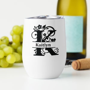 Könnte beinhalten: Ein weißer Edelstahl-Weinglas mit einem schwarzen floralen Design und dem Buchstaben K. Der Name Kaitlyn ist in Schwarz unter dem Design geschrieben.