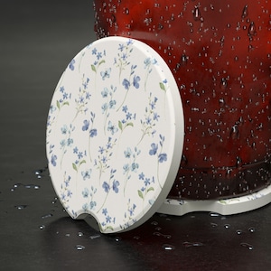 May include: A white ceramic coaster with a blue floral pattern. The coaster has a cutout for easy gripping.