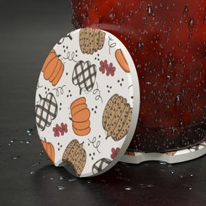 May include: A white ceramic coaster with a fall-themed design featuring pumpkins, plaid, and polka dots. The coaster is round and has a slightly raised edge.