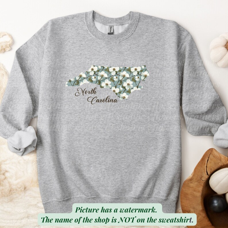 May include: A light gray sweatshirt featuring a floral design in the shape of North Carolina. The design is filled with white flowers on a teal background, with the words "North Carolina" in a script font.