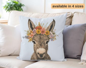 May include: A throw pillow with a light blue background featuring a watercolor illustration of a donkey wearing a flower crown. The donkey has brown fur and a sweet expression. The flowers in the crown are a mix of yellow, orange, pink, and purple.