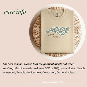 May include: A tan sweatshirt with a floral North Carolina design. The state outline is filled with white flowers and green leaves, with "North Carolina" in script below. The shirt is folded on a woven mat.