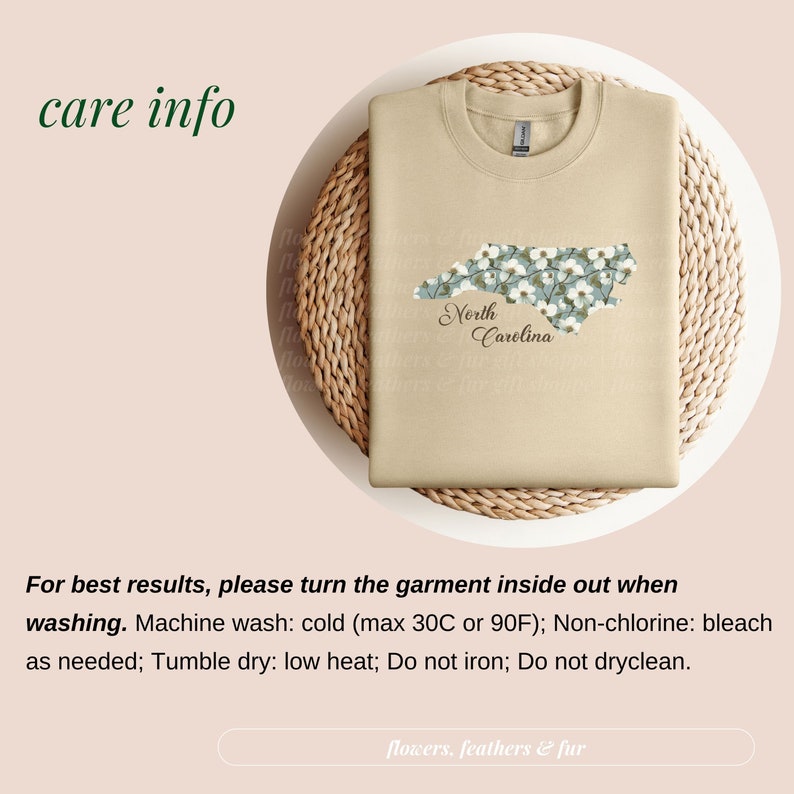 May include: A tan sweatshirt with a floral North Carolina design. The state outline is filled with white flowers and green leaves, with "North Carolina" in script below. The shirt is folded on a woven mat.