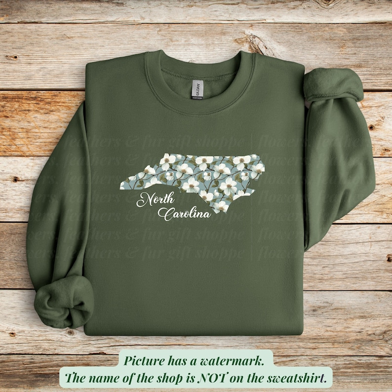 May include: An olive green sweatshirt featuring a floral North Carolina state outline. The state shape is filled with white flowers on a light blue background, with "North Carolina" in script below.
