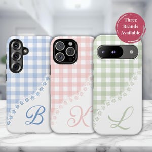 May include: Three phone cases in pastel shades of blue, pink, and green. Each case has a gingham pattern, a white floral lace design, and a monogram initial. The cases are for different phone models. The text "Three Brands Available" is in a circle.