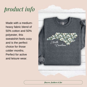 May include: A dark gray sweatshirt featuring a floral North Carolina state outline. The design includes white flowers on a light blue background, with "North Carolina" in script. The sweatshirt is made of a medium-heavy fabric blend.