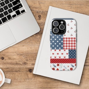 May include: A patchwork-style phone case featuring a patriotic design. The case incorporates blue and white stars, red and white gingham, red bows, and floral patterns. The case is on a white book, with a laptop and coffee cup in the background.