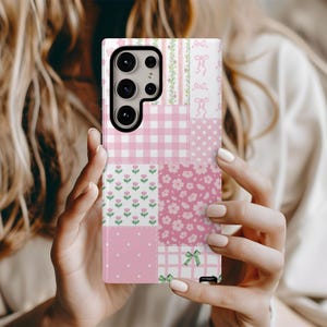 May include: A pink and white patchwork phone case featuring various patterns, including gingham, floral, and polka dots. The case has a rectangular shape and is designed to fit a smartphone. The case is held by a person.