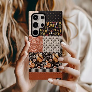 以下が含まれることがあります： パッチワークデザインの模様入り携帯電話ケース。ケースには、茶色、黒、オレンジの色合いのフローラル、キツネ、幾何学模様のセクションが含まれています。ケースにはカメラの切り抜きがあります。