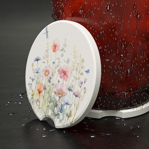 May include: A white ceramic coaster with a watercolor design of pink, blue, and yellow wildflowers. The coaster has a cutout on the bottom to fit around a glass.