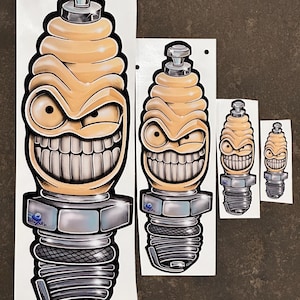 以下が含まれることがあります： いたずらっぽい笑顔をした漫画のスパークプラグが描かれたステッカーが4つの異なるサイズで用意されています。スパークプラグは黄色とグレーで、銀色のナットが付いています。ステッカーは白い背景です。