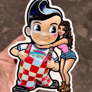 Puede incluir: Una pegatina con un personaje de dibujos animados que lleva un mono rojo y blanco a cuadros con el texto "WAK'OUT" en la parte delantera. El personaje sostiene un pincel y está siendo abrazado por una mujer de dibujos animados que lleva una camisa rosa y pantalones cortos de mezclilla.