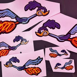 May include: A selection of Road Runner cartoon character stickers. The stickers depict the Road Runner in full flight, with blue wings, purple head feathers, and a yellow beak. The stickers have a red and orange flame trail.