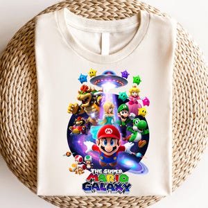 Może przedstawiać: Kremowa koszulka z kolorową grafiką Mario i innych postaci z gry Super Mario Galaxy. Projekt zawiera statek kosmiczny obcych, gwiazdy i logo gry w kolorze niebieskim, czerwonym i zielonym.
