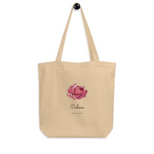 May include: Beige tote bag with a pink and red floral design. The word "Calma" is written in cursive below the flower, with the phrase "SIMPLE MOM & KIDS" underneath. The bag has long straps and a black handle.