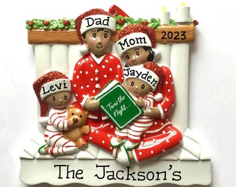 May include: A personalized Christmas ornament featuring a family of four in red and white pajamas, reading a book together in front of a fireplace. The ornament is decorated with the names "Dad", "Mom", "Jayden", and "Levi", and the year "2023". The text "The Jackson's" is written on the bottom of the ornament.