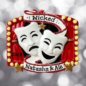 May include: A red and yellow Christmas ornament with two white masks, one smiling and one frowning. The ornament has the words "Wicked" and "Natasha & Ale" on it.