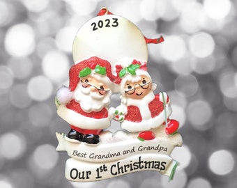 Puede incluir: Adorno navideño blanco con una figura de Papá Noel y la Sra. Claus con un círculo en blanco para una foto. El adorno tiene una pancarta que dice "Mejor abuela y mejor abuelo Nuestra primera Navidad" y el año 2023.